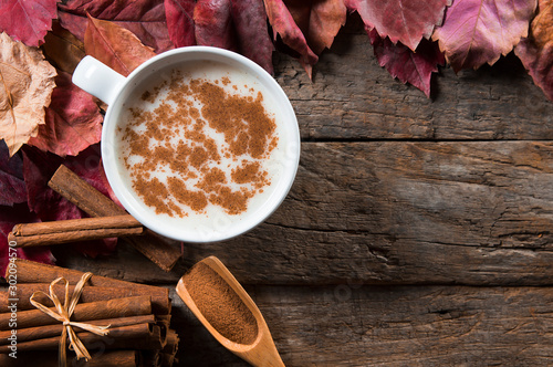 Top view white cup of salep milky hot drink of Turkey with cinnamon sticks, powder and autumn leaves on wooden backdrop photo