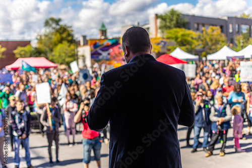 Selective focus of man on stage from the back taking to crowd. Group of people at open public space with banners listening to lider at background.