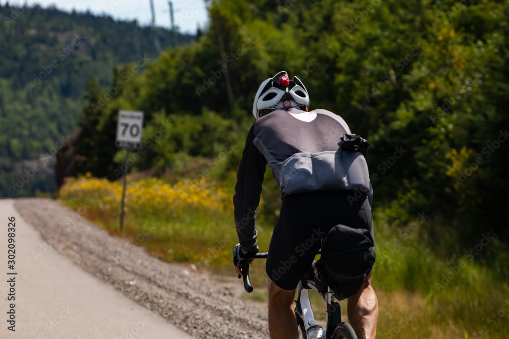 Selective focus on cyclist viewed from the back while cycling a mountain highway with sport clothing on. Forest environment, woods in the background Selective focus on cyclist viewed from the back while cycling a mountain highway with sport clothing on. Forest environment, woods in the background