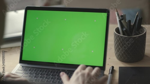 Over the shoulder shot of young woman working on modern laptop with green screen mock-up. Hands on keyboard.