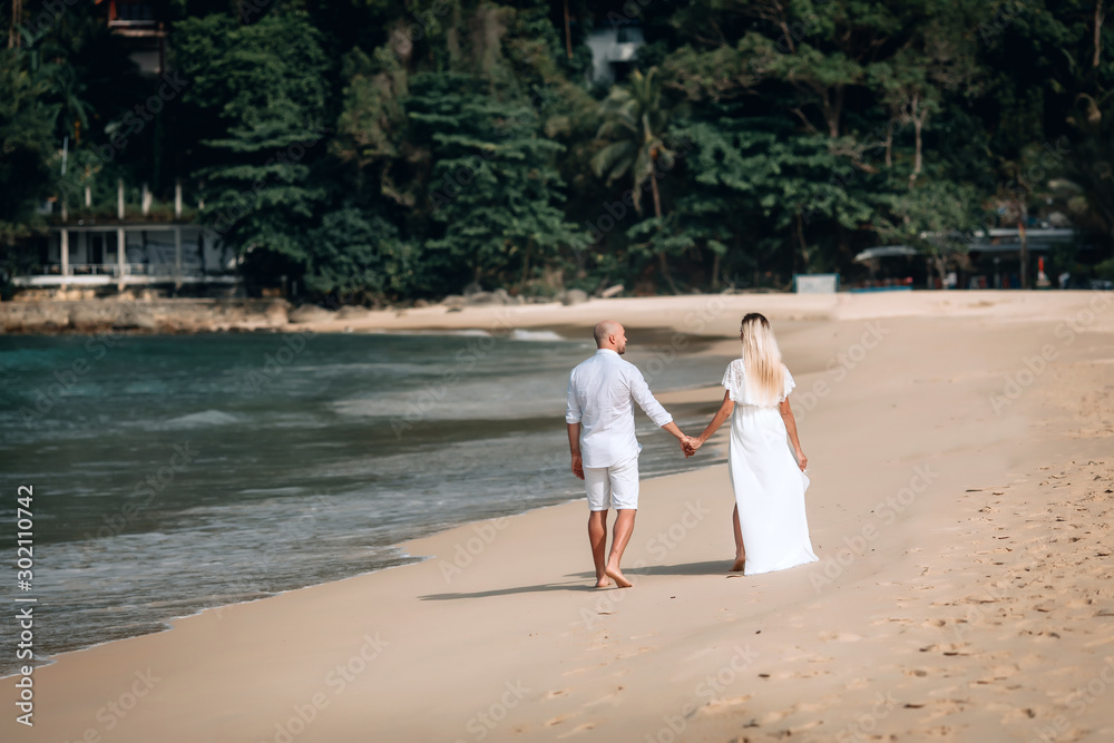 Attractive trendy young couple in white clothes are walking along on the beach and holding hands. Beautiful blonde with long hair and a bald man are on vacation. Phuket. Thailand.