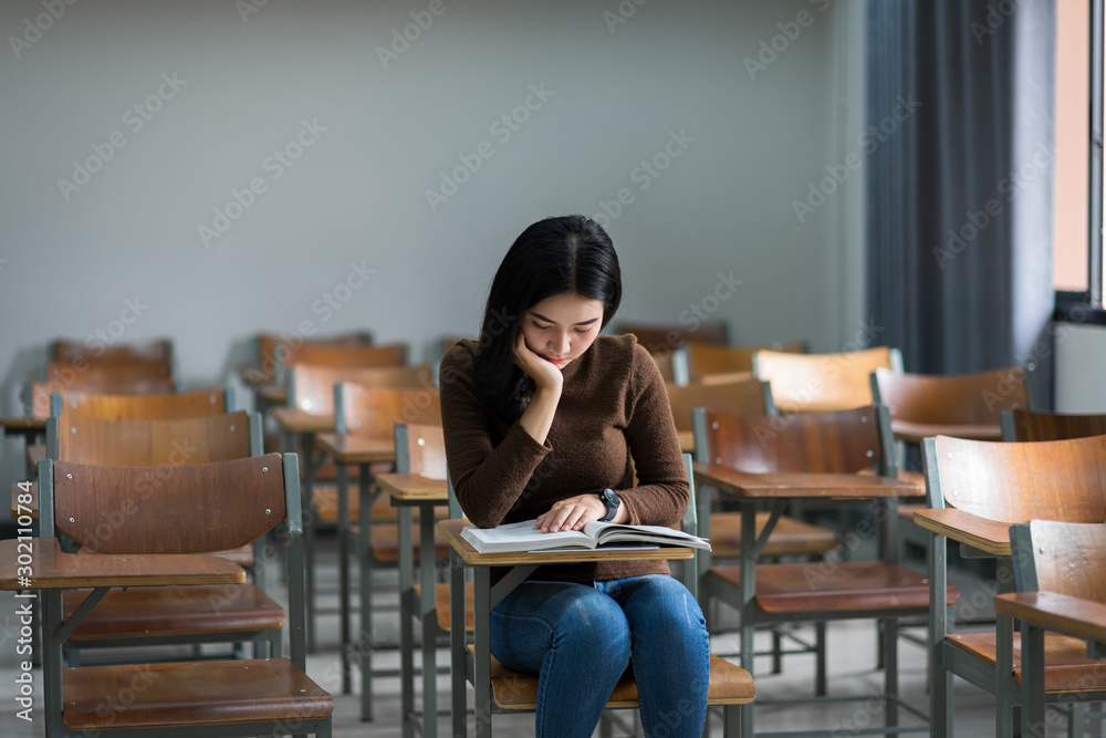 A teenager woman university student sits on wooden a chair concentrates reading textbook in the classroom preparing for examination.