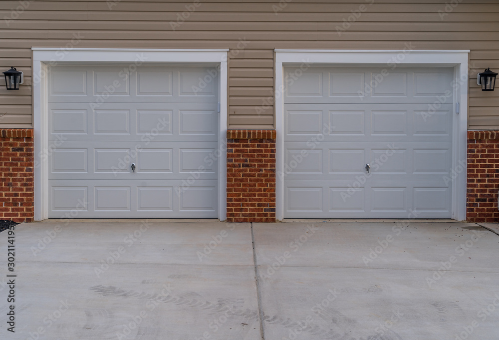 White electronic garage doors on a double garage driveway with beige horizontal lap vinyl siding and red brick accent on a new construction luxury American single family home in the East Coast USA