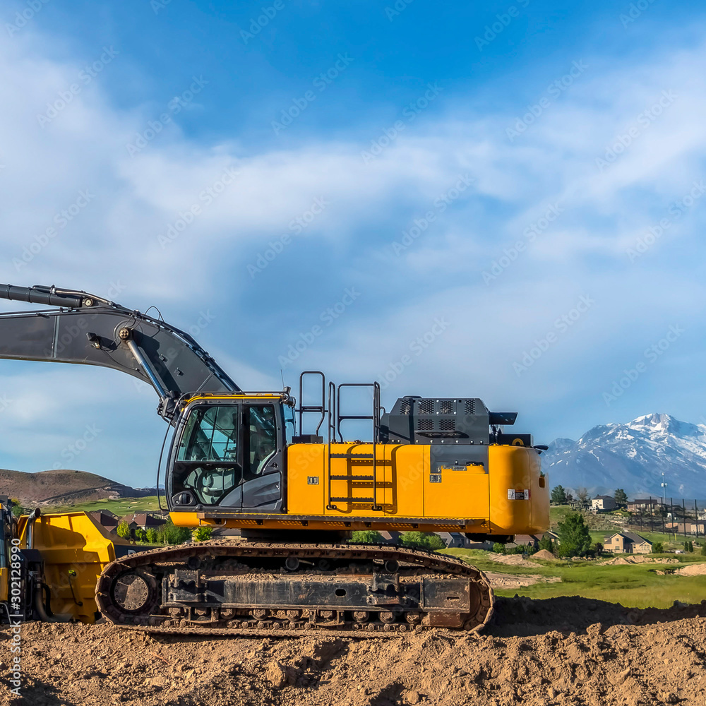 Square frame Excavator at a construction site with mountain homes and cloudy sky background Square frame Excavator at a construction site with mountain homes and cloudy sky background