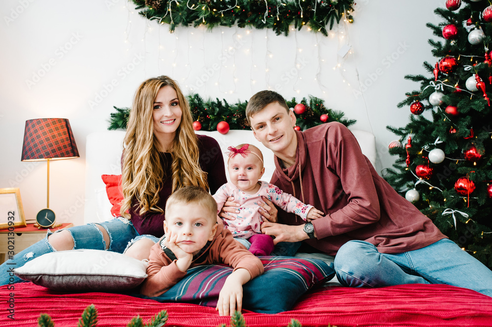 The dad, mom hug little son and daughter near Christmas tree. Happy New Year and Merry Christmas. Christmas decorated interior. The concept of family holiday. Close up. The dad, mom hug little son and daughter near Christmas tree. Happy New Year and Merry Christmas. Christmas decorated interior. The concept of family holiday. Close up.
