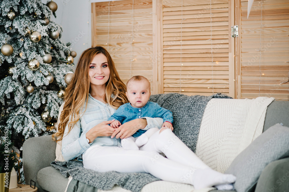 Baby girl, daughter with mom on sofa near Christmas tree. Happy New Year and Merry Christmas. Christmas decorated interior. The concept of family holiday.