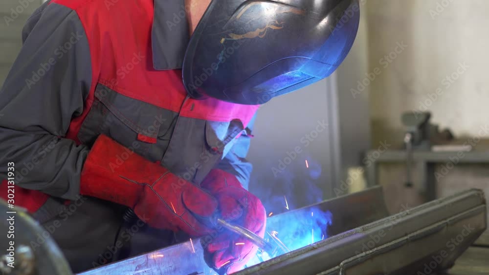 Man in gloves and welding helmet works with metal, high temperature welding, argon welding, sparks, blue glow, hard work, factory production, employee of welding workshop at work, complex product Man in gloves and welding helmet works with metal, high temperature welding, argon welding, sparks, blue glow, hard work, factory production, employee of welding workshop at work, complex product