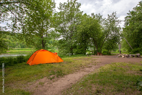An orange double tent stands in a clearing in a forest by the river. The sky is gray with clouds.