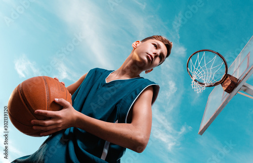 Sports and basketball. A young teenager in a black tracksuit stands with a ball in his hands and prepares to throw the ball into the ring. Blue sky. Bottom view © _KUBE_