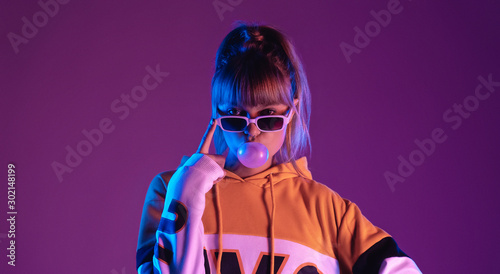Pretty young 20s fashion teen girl model wear glasses blowing bubble gum looking at camera standing at purple studio background, igen teenager in trendy stylish night glow 80s 90s concept, portrait