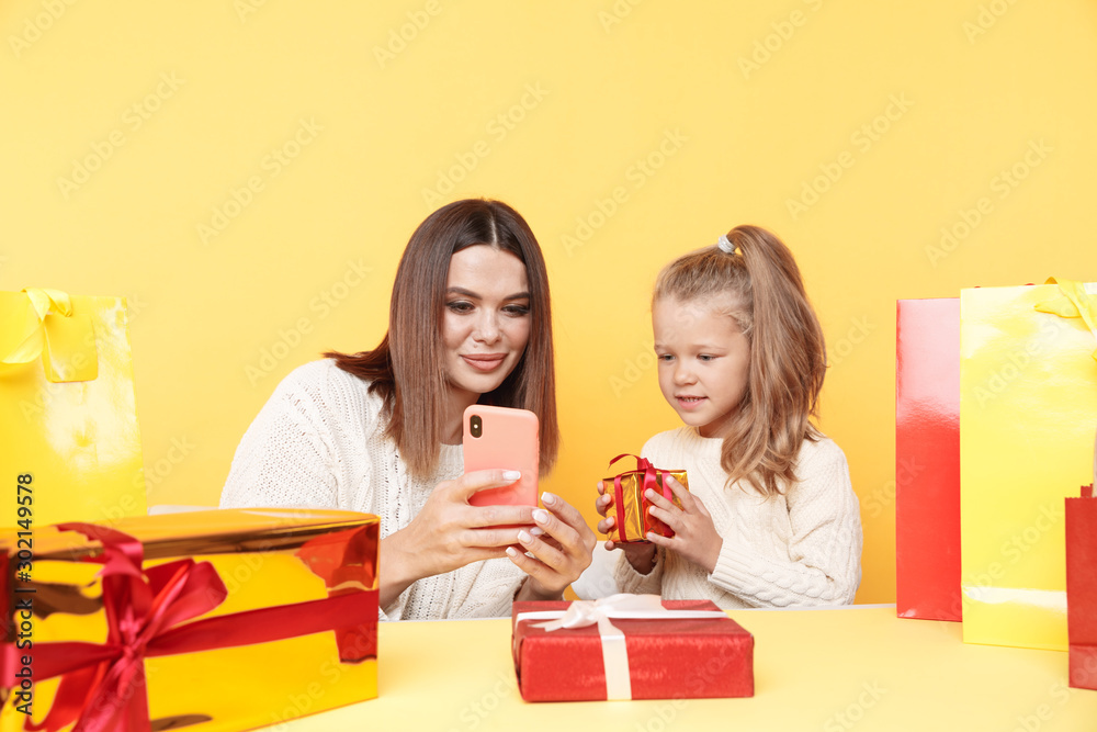 Pretty mother with little daughter sitting at the desk with gifts and using phone to choose presents online