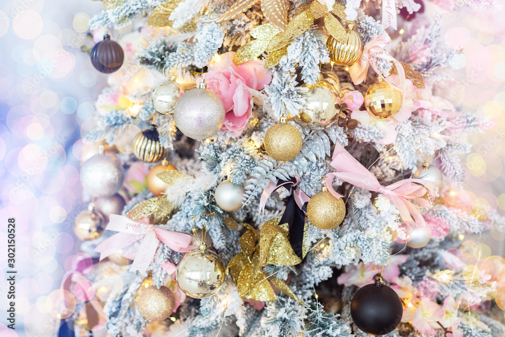 Obraz premium Close-up shiny pink gold decorated Christmas tree with lights ribbons, baubles and balls at house indoors. Xmas decoration background. Winter holidays card. Toned with bright bokeh lights