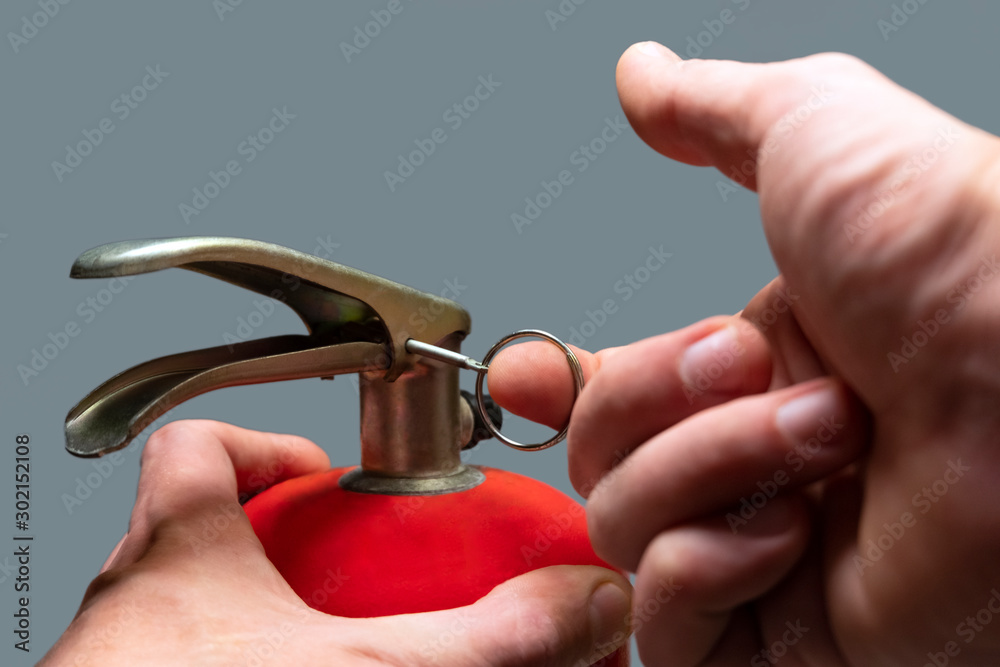A man pulls out a safety ring from a red fire extinguisher. Close-up. Male  finger pulls out a pin. Fire fighting regulations. Emergency Prevention.  Shallow depth of field. Selective focus.Isolate gray Stock-Foto