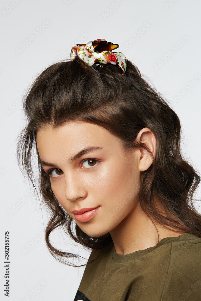 Naklejka premium Closeup half-turn face portrait of girl with dark wavy hair, wearing khaki t-shirt. Her hair is pulling with brown hairpin crab with white bow with red flower ornament. 