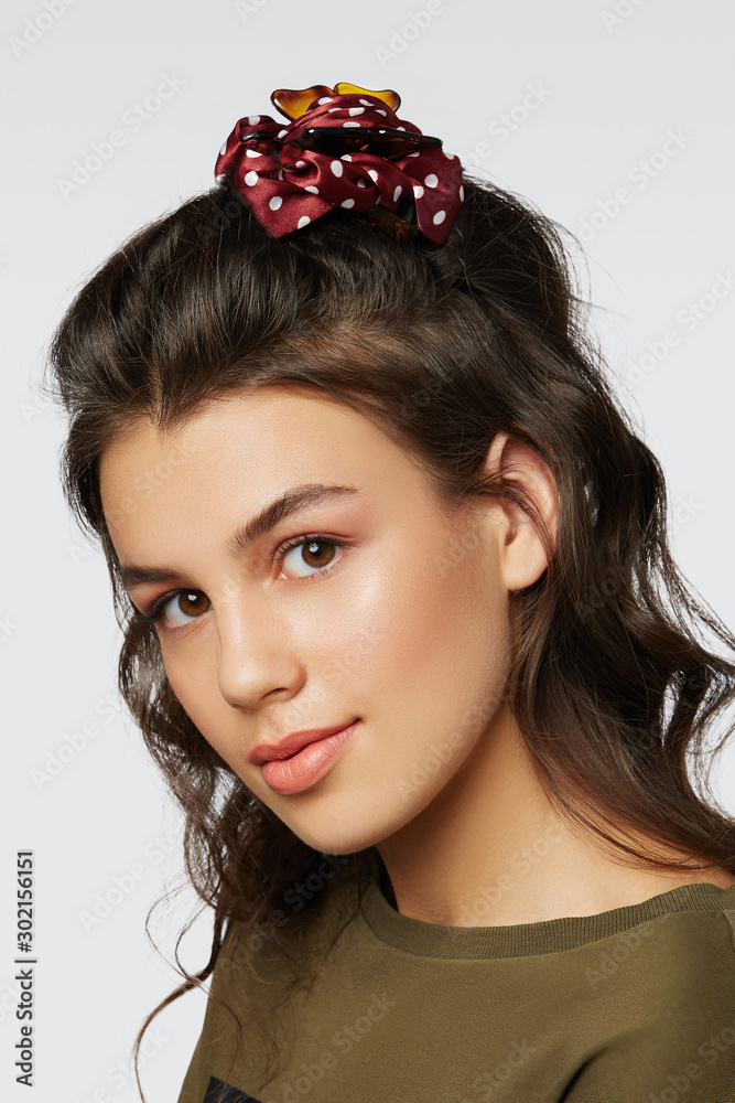 Naklejka premium Closeup half-turn face portrait of girl with dark wavy hair, wearing khaki t-shirt. Her hair is pulling with brown hairpin crab with red polka dot bow. Girl is looking straight and smiling.