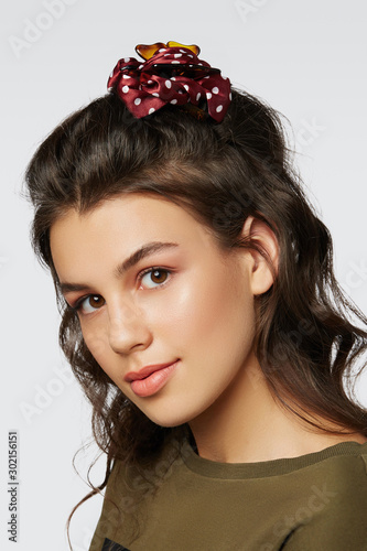 Closeup half-turn face portrait of girl with dark wavy hair, wearing khaki t-shirt. Her hair is pulling with brown hairpin crab with red polka dot bow. Girl is looking straight and smiling.