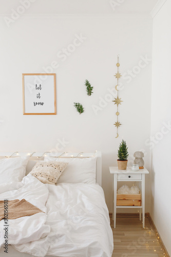 New year winter home interior decor. Holiday decorated room. Stylish cozy white scandinavian bedroom: bed, little christmas tree in a pot on bedside table, pine branches, frame with text LET IT SNOW.