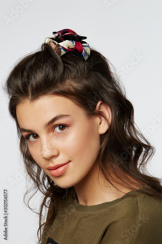 Closeup half-turn face portrait of girl with dark wavy hair, wearing khaki t-shirt. Her hair is pulling with black hairpin crab, decorating with white patterned bow. 