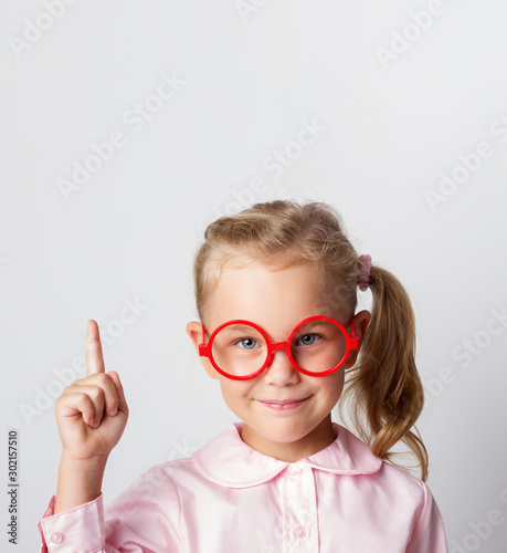 Close-up portrait of amazed pretty young girl schoolgirl in red glasses and a light blouse  pointing finger up  looking at camera with a smile  isolated on a light background