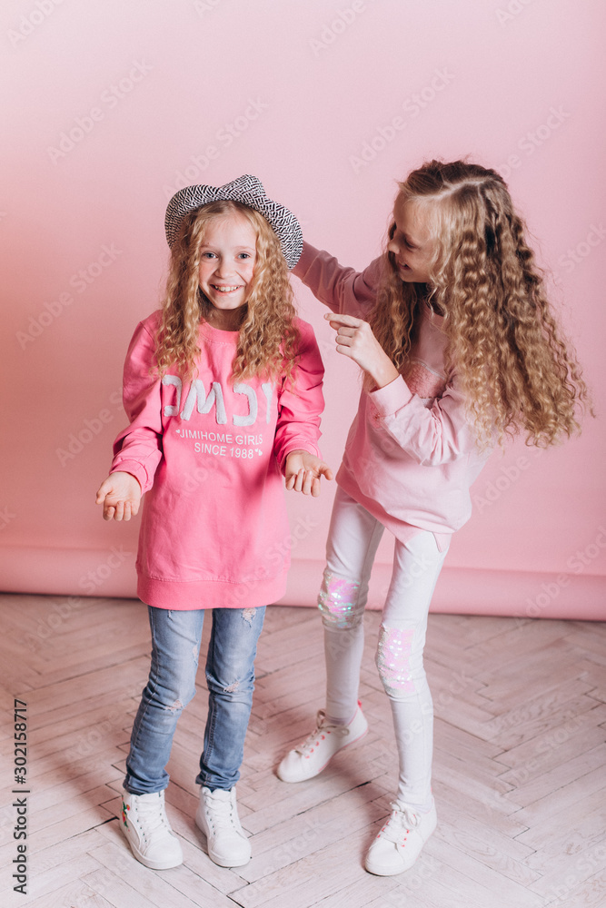 Close up portrait of young cute adorable caucasian sisters with curly hairstyle on soft pink background indoors in studio. Positive leisure concept, childhood happiness concept. Close up portrait of young cute adorable caucasian sisters with curly hairstyle on soft pink background indoors in studio. Positive leisure concept, childhood happiness concept.