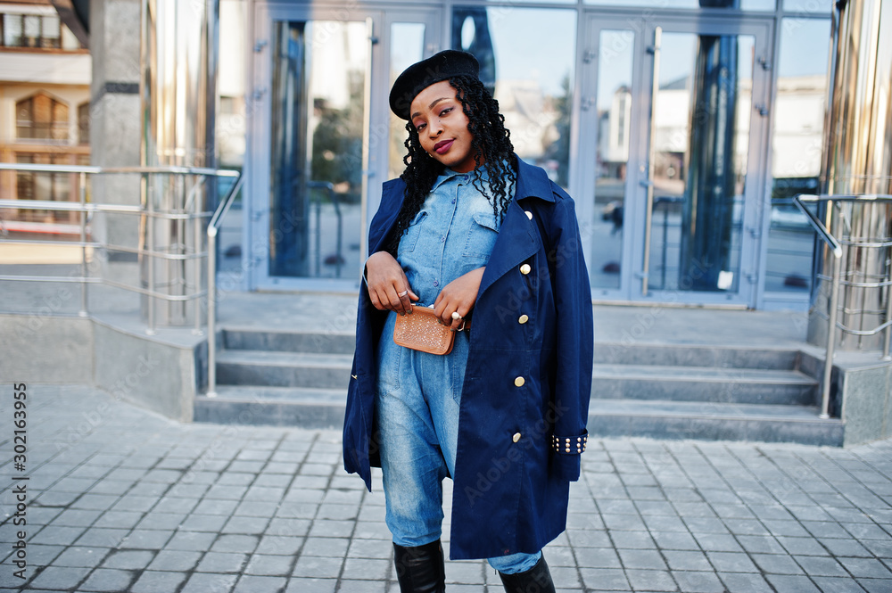 Stylish fashionable african american women in jeans wear and black beret with blue coat against modern building.