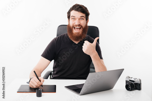 Photo of smiling bearded man showing thumbs up standing at office table, freelance phtography job photo