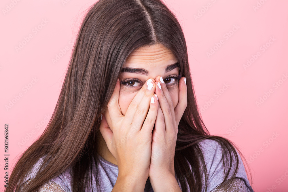 Photo of frightened woman with brown hair in basic t-shirt keeping hands over her face and looking at camera isolated over pink background