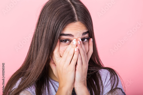 Photo of frightened woman with brown hair in basic t-shirt keeping hands over her face and looking at camera isolated over pink background