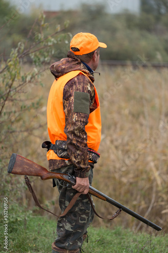 A man with a gun in his hands and an orange vest on a pheasant hunt in a wooded area in cloudy weather. Hunter with dogs in search of game.