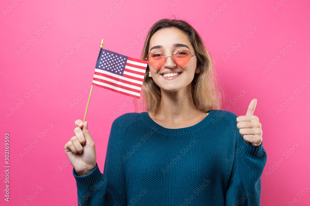 Happy young woman heart-shaped glasses holding American flag against a studio pink background Happy young woman heart-shaped glasses holding American flag against a studio pink background