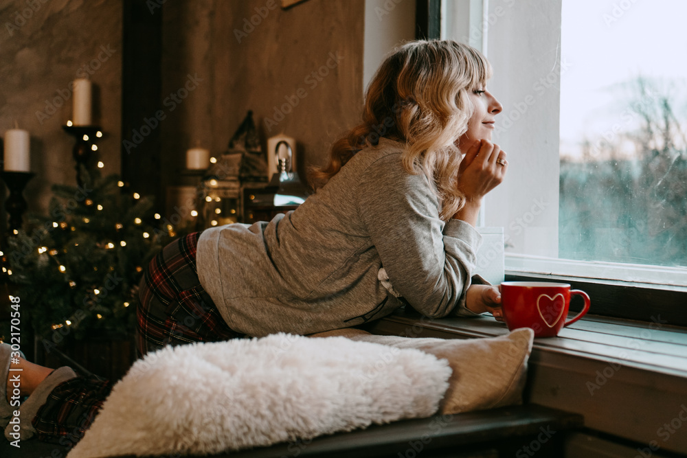 Young woman in plaid with cup of hot tea in a Christmas cozy interior. The concept of preparation for the holidays, Make a wish and dream - obrazy, fototapety, plakaty 