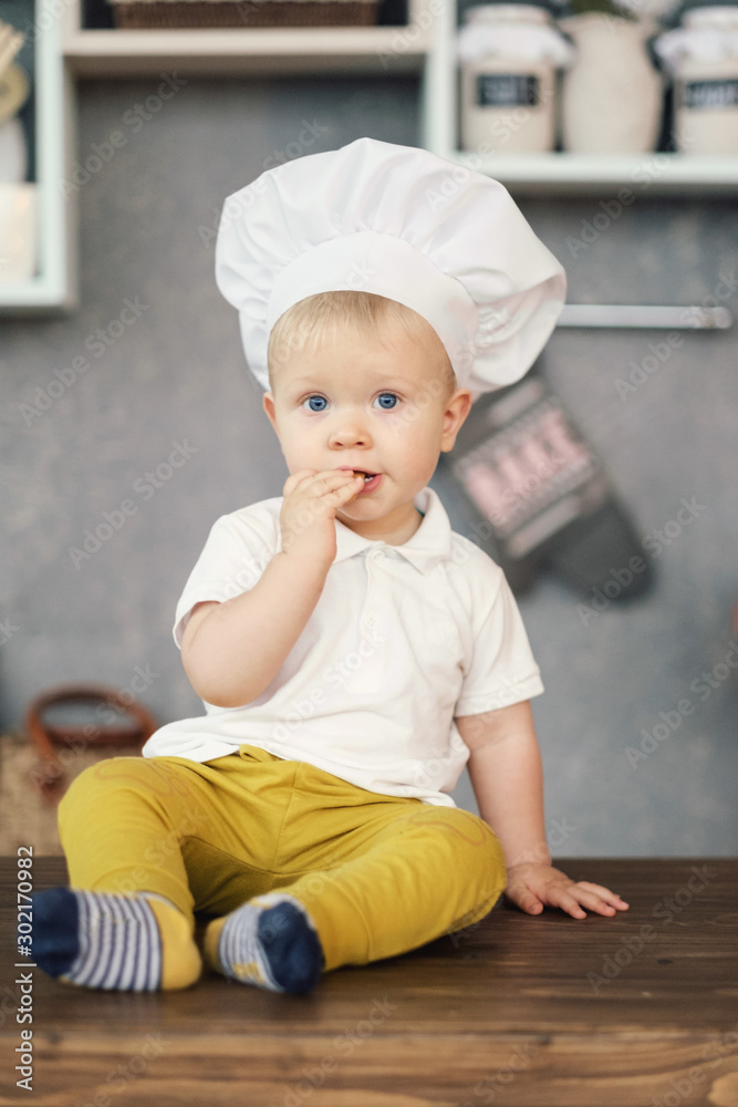 one little boy in hat of chef in kitchen sitting on the table and eat one little boy in hat of chef in kitchen sitting on the table and eat