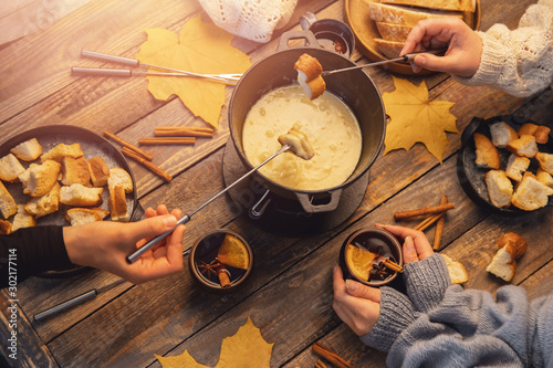 Meeting of friends, girl is cooking Swiss fondue assorted cheeses dinner on fire, hands holding mulled wine background wood board, top view photo