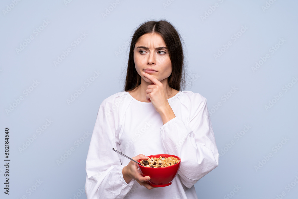 Young woman over isolated blue background holding a bowl of cereals and thinking Young woman over isolated blue background holding a bowl of cereals and thinking