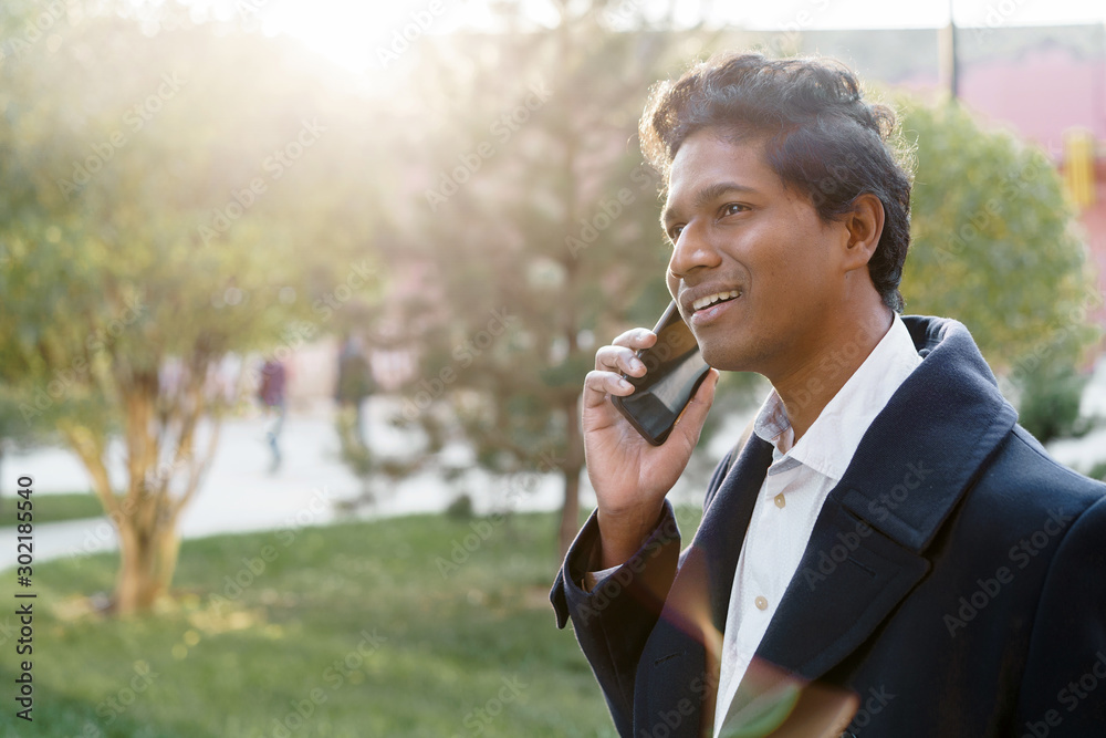 Attractive handsome Indian man in a business suit and coat with a backpack walking on a sunny autumn day in city park and talking on the phone with friend about meeting, looking back, smiling Attractive handsome Indian man in a business suit and coat with a backpack walking on a sunny autumn day in city park and talking on the phone with friend about meeting, looking back, smiling