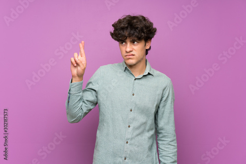 Young man over isolated purple wall with fingers crossing and wishing the best
