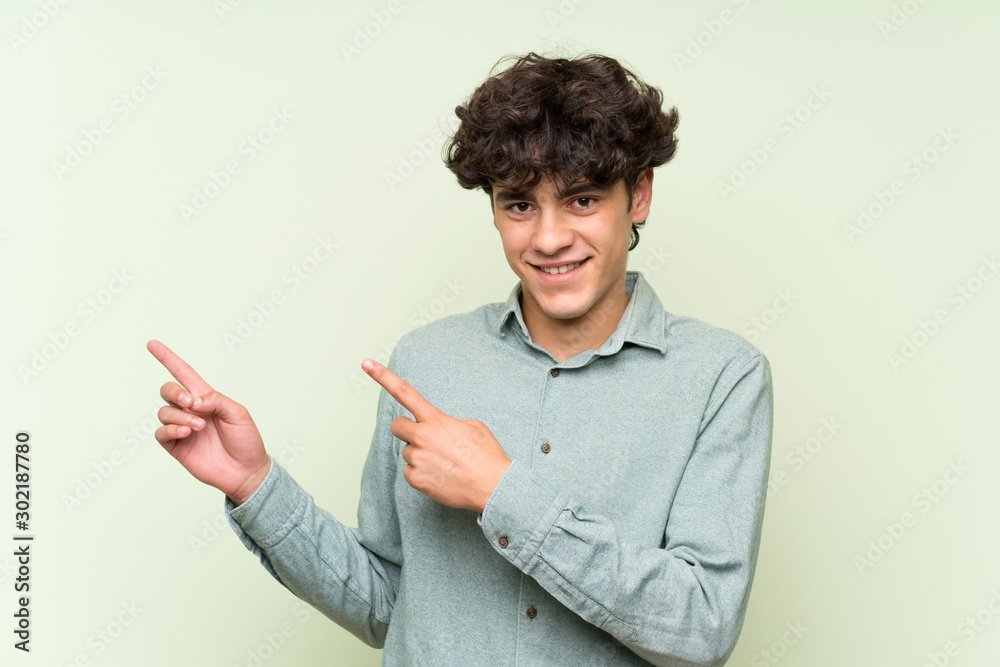 Young man over isolated green wall pointing finger to the side Young man over isolated green wall pointing finger to the side