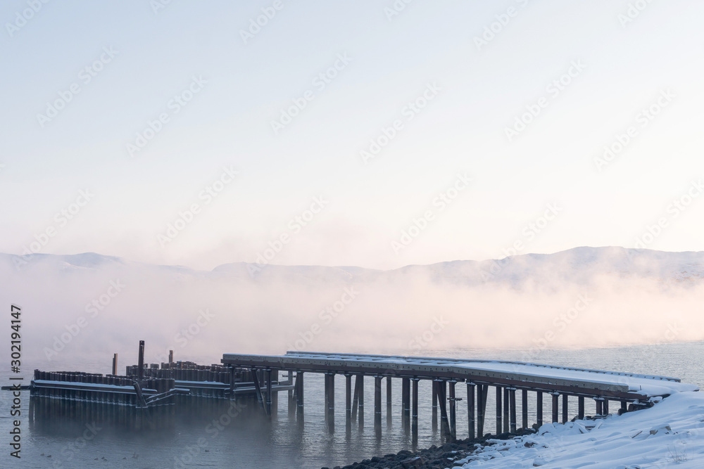Obraz premium old wooden pier for large ships on the river bank covered in thick white fog from the water at dawn on a frosty winter day outside the Arctic Circle