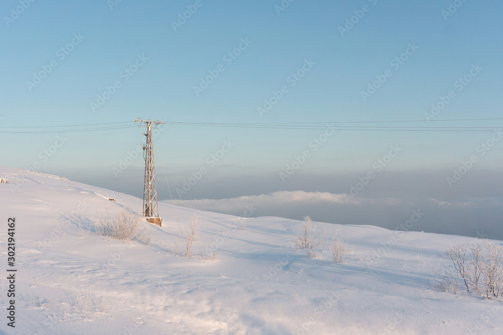 a metal tower holding wires stands on a hill from where the bottom is not visible due to fog during severe frosts beyond the Arctic Circle in the far north