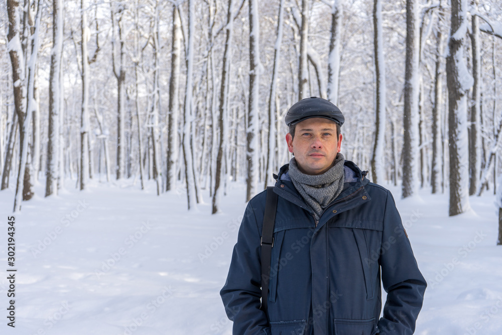 Portrait of handsome middle-aged man walking in winter snowy park or forest. Attractive man in jacket, scarf and cap looking at camera. Winter mood, authentic lifestyle concept, stylish male outfit
