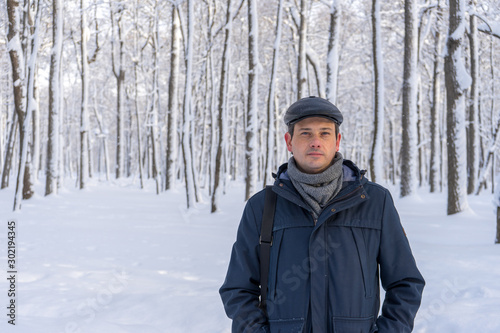 Portrait of handsome middle-aged man walking in winter snowy park or forest. Attractive man in jacket, scarf and cap looking at camera. Winter mood, authentic lifestyle concept, stylish male outfit