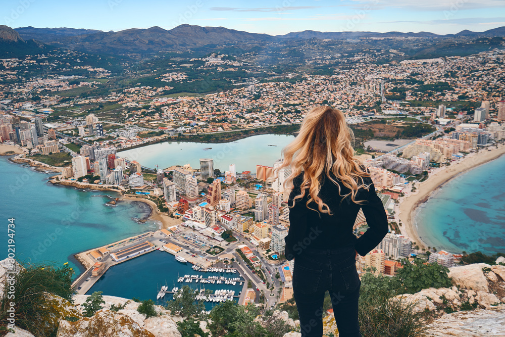 Rear view woman climbed up on peak of Penon de Ifach rock enjoy picturesque view Mediterranean Sea, mountain range, cityscape scenery. Tourism, famous beautiful place visit hiking concept. Calpe Spain