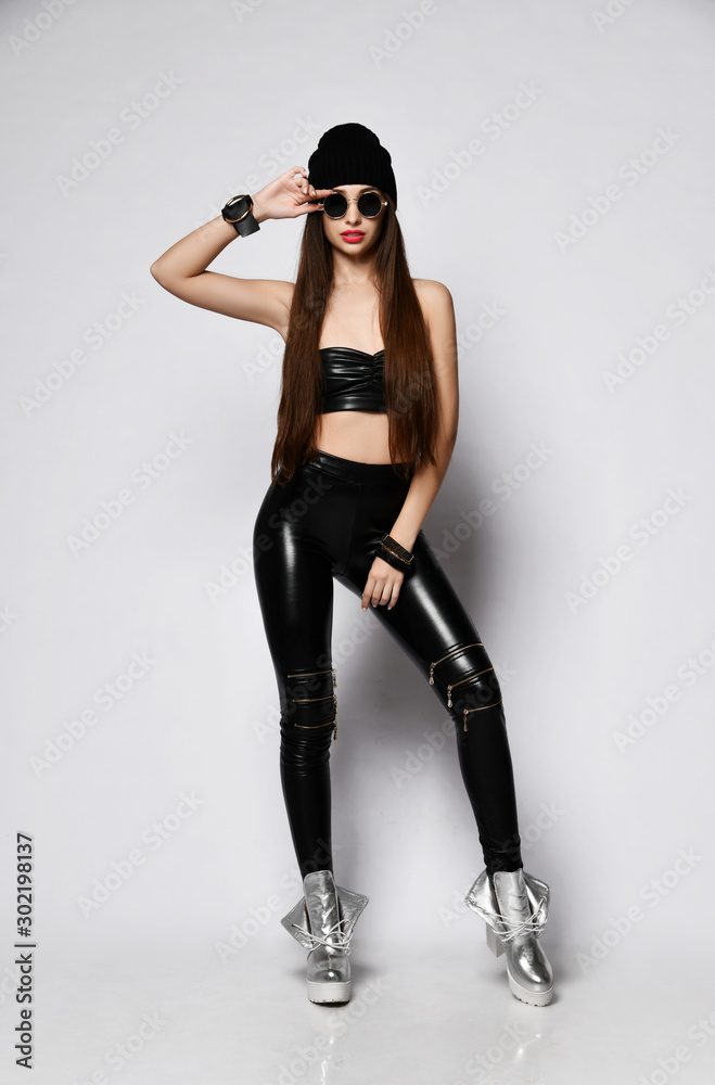 Brutal grunge girl in black hat, tight leather leggings with zippers is  fashion posing holding her round sunglasses foto de Stock | Adobe Stock