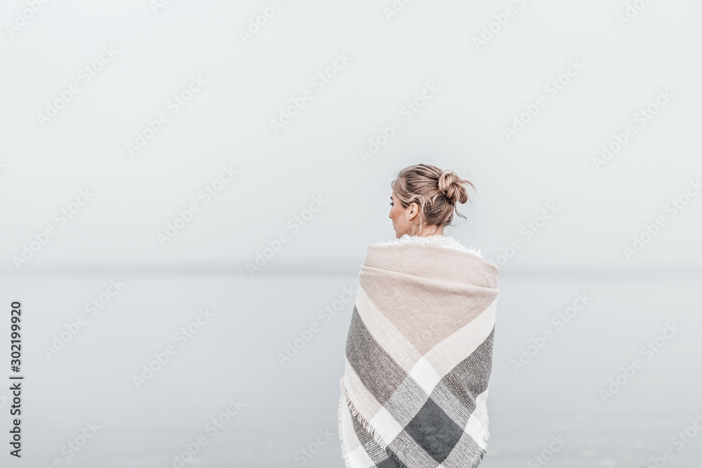 Pretty blonde woman wrapped in warm blanket standing on the cold autumn beach. - obrazy, fototapety, plakaty 