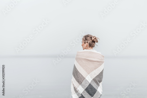 Pretty blonde woman wrapped in warm blanket standing on the cold autumn beach.