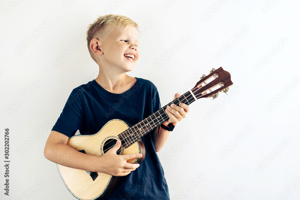 Young blond teenager plays ukulele and sings a fun song. Portrait of a laughing child engaged in a favorite hobby on a big background with place for text. Young blond teenager plays ukulele and sings a fun song. Portrait of a laughing child engaged in a favorite hobby on a big background with place for text.