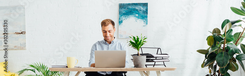 Smiling freelancer using laptop at desk in living room, panoramic shot