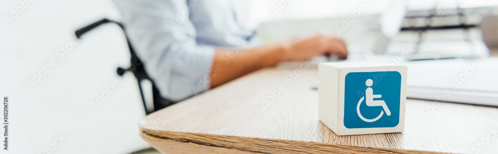 Selective focus of cube with disabled sign and man in wheelchair sitting at table, panoramic shot Selective focus of cube with disabled sign and man in wheelchair sitting at table, panoramic shot