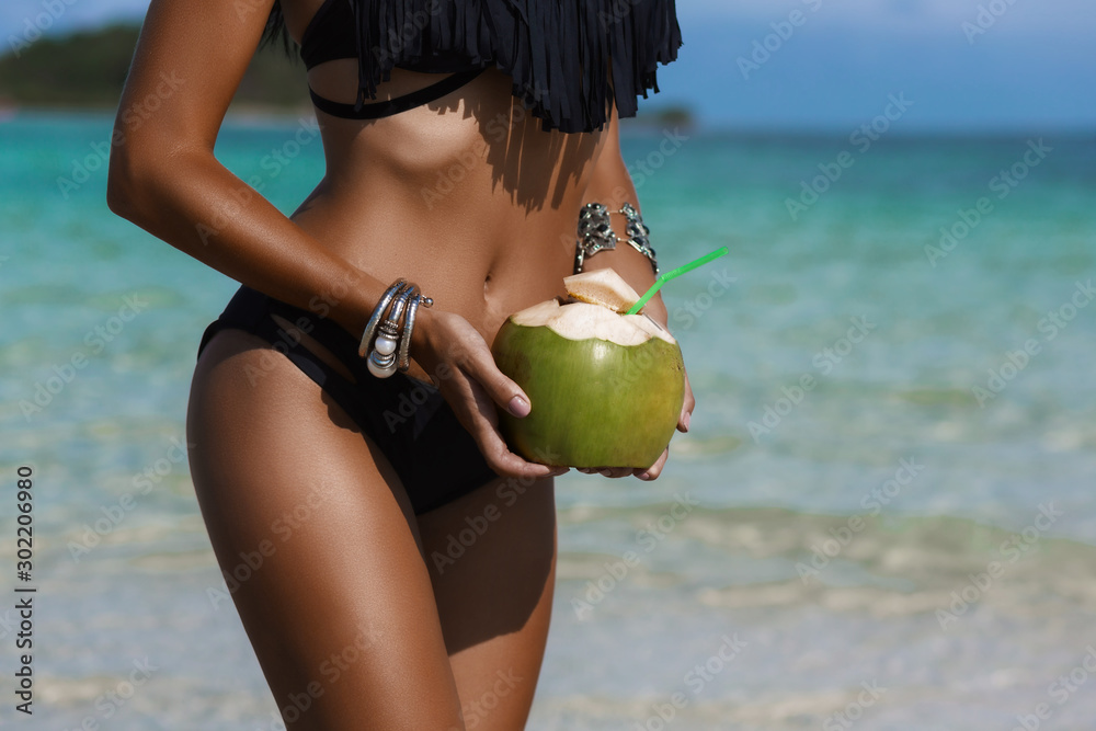 Beautiful tanned sexy lady, a perfect body, holding a fresh coconut, against the background of a tropical island, on the beach, vacation travel, happiness and relaxation - obrazy, fototapety, plakaty 