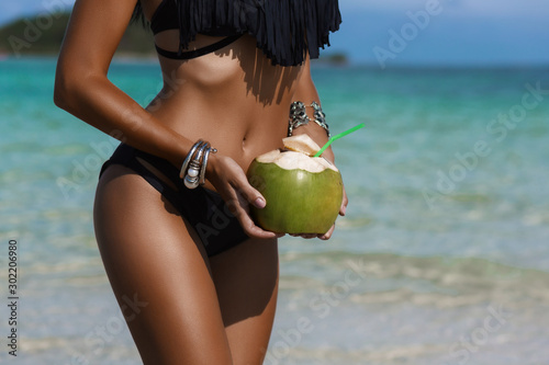 Beautiful tanned sexy lady, a perfect body, holding a fresh coconut, against the background of a tropical island, on the beach, vacation travel, happiness and relaxation